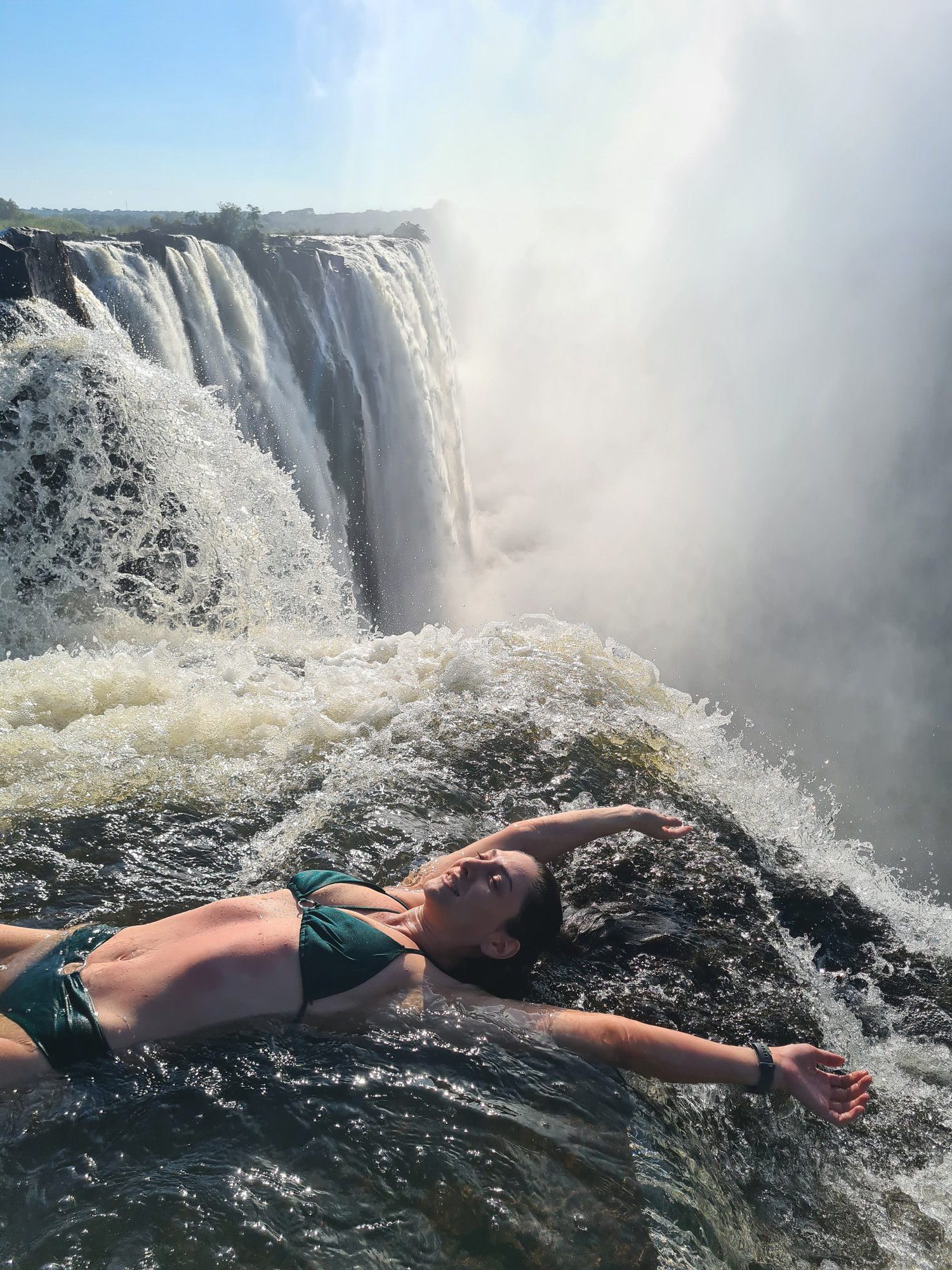 Kelly with arms raised in the Victoria Falls infinity pool, mist rising from the gorge